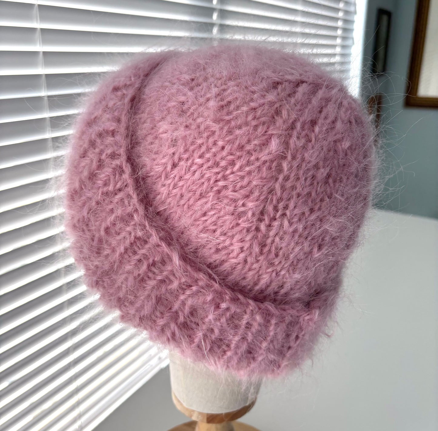 Pink knitted hat on a mannequin head against a window with blinds.