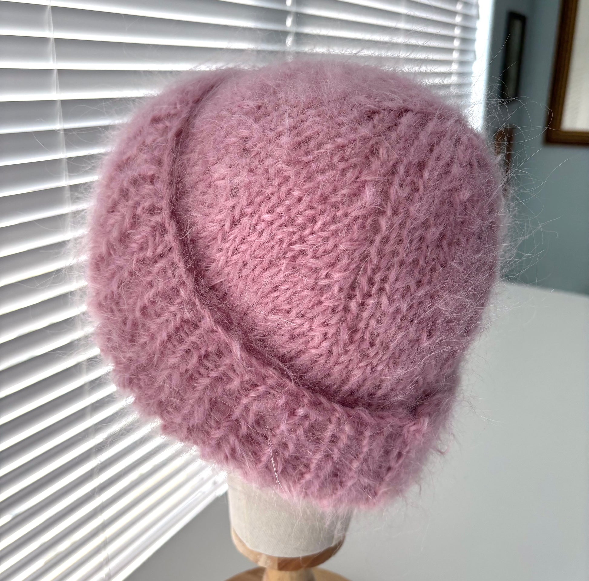 Pink knitted hat on a mannequin head against a window with blinds.